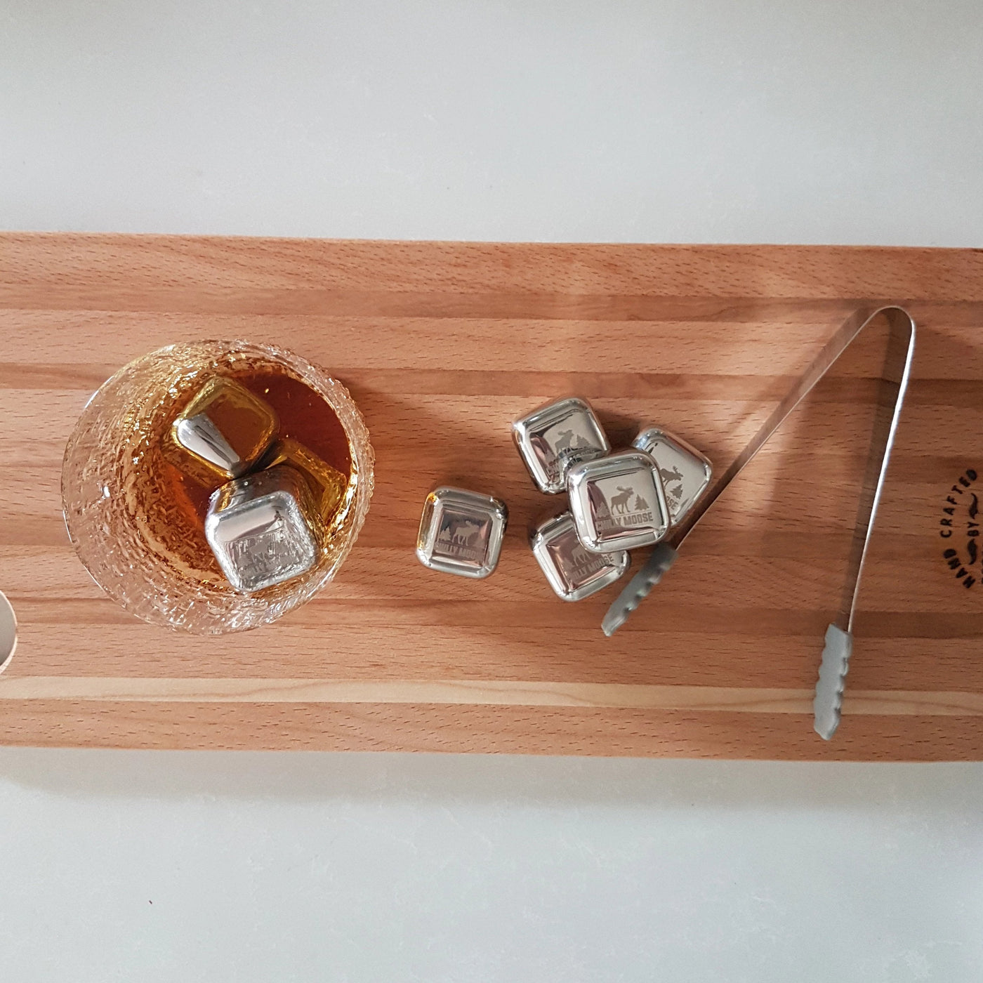Three Chilly Moose stainless steel reusable ice cubes in a cocktail glass with the remainder stacked beside with included tongs, displayed on a wooden cutting board